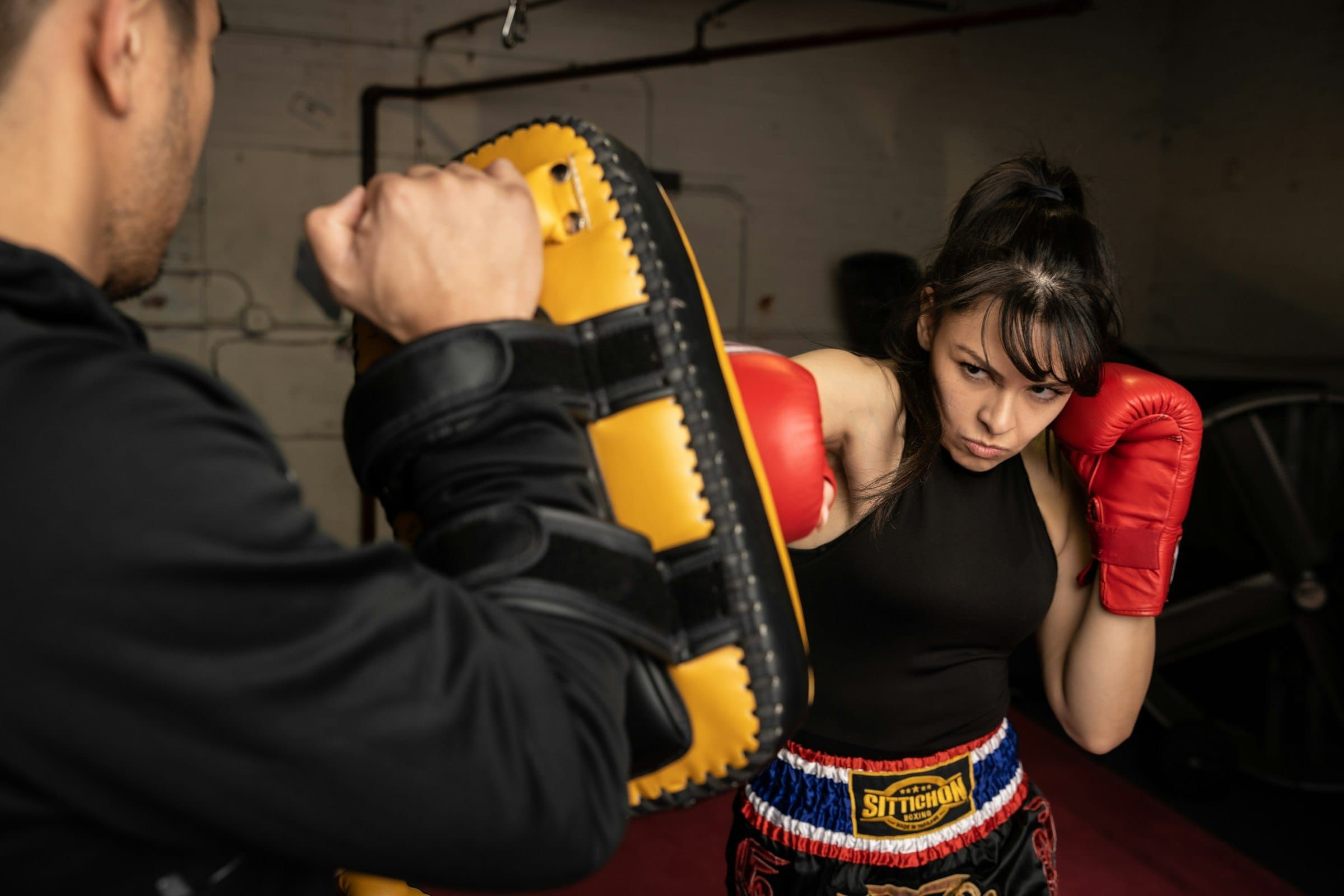 A woman in a black tank top and red boxing gloves hitting a pad held by a man in a dim gym.