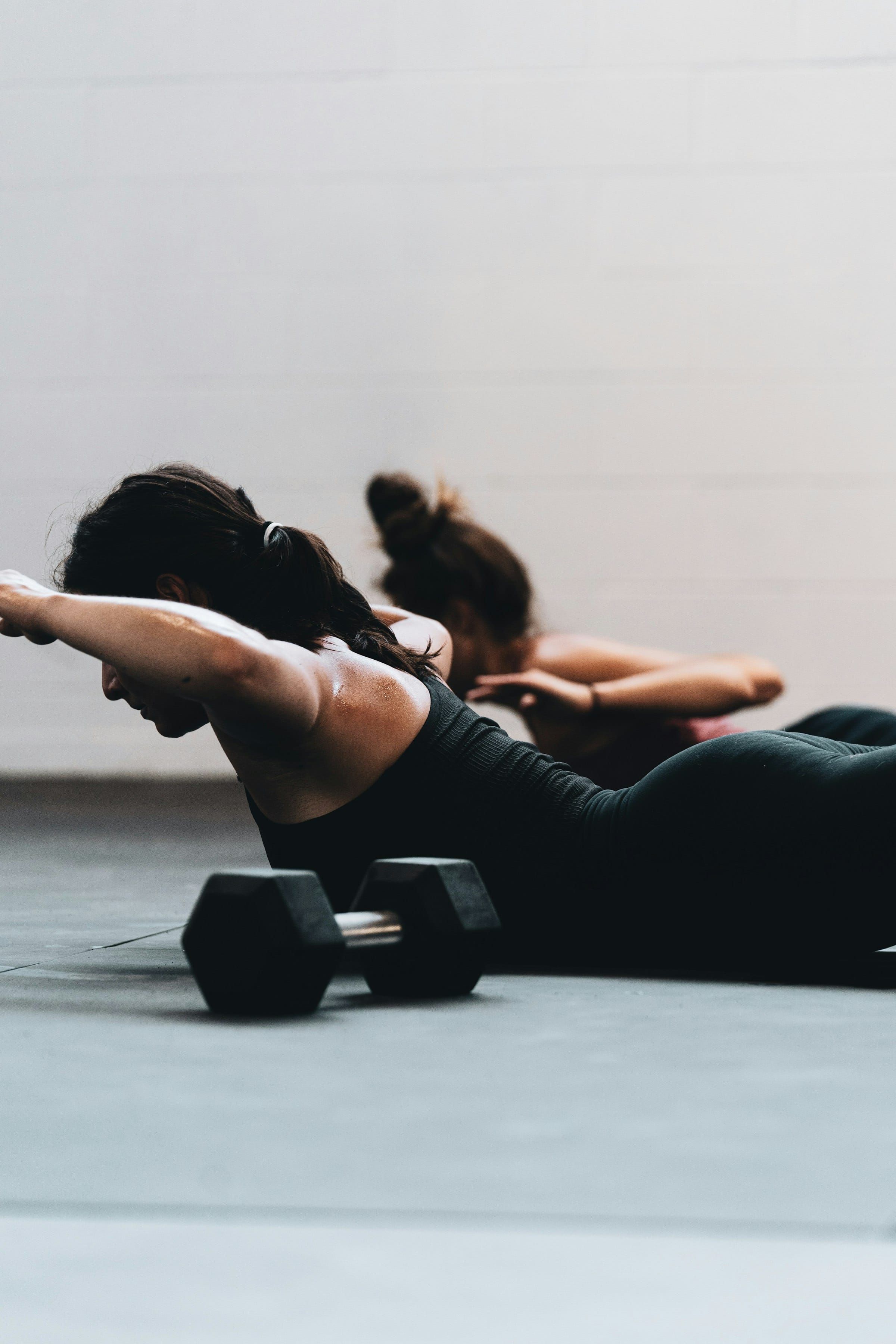 Two women lying on their stomachs on the floor doing back extensions, a black dumbbell on the floor in front of them.