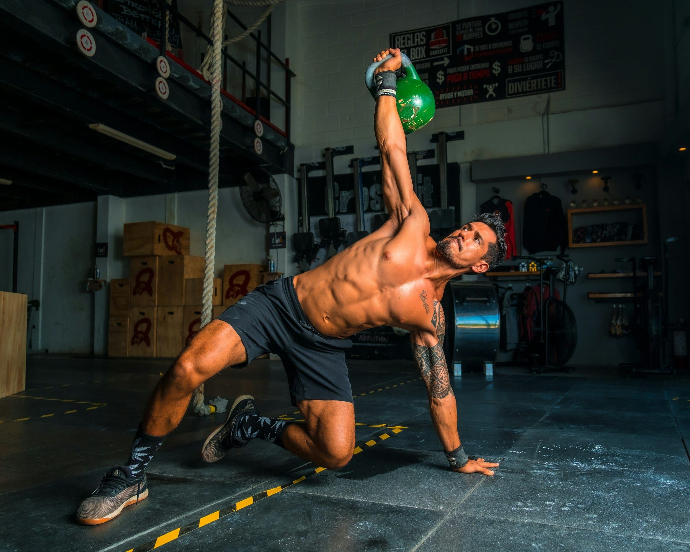 A shirtless man in black shorts doing a kettlebell get-up, a green kettlebell held overhead, in a dim gym.