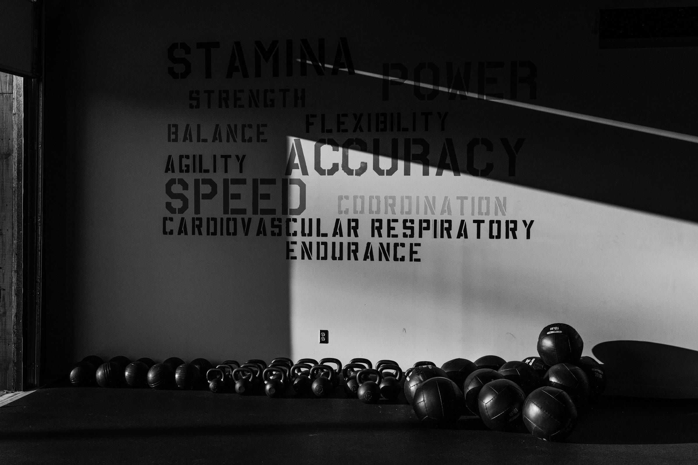 Kettlebells and medicine balls lined up on a gym floor in front of a wall lettered with fitness words, in black and white.