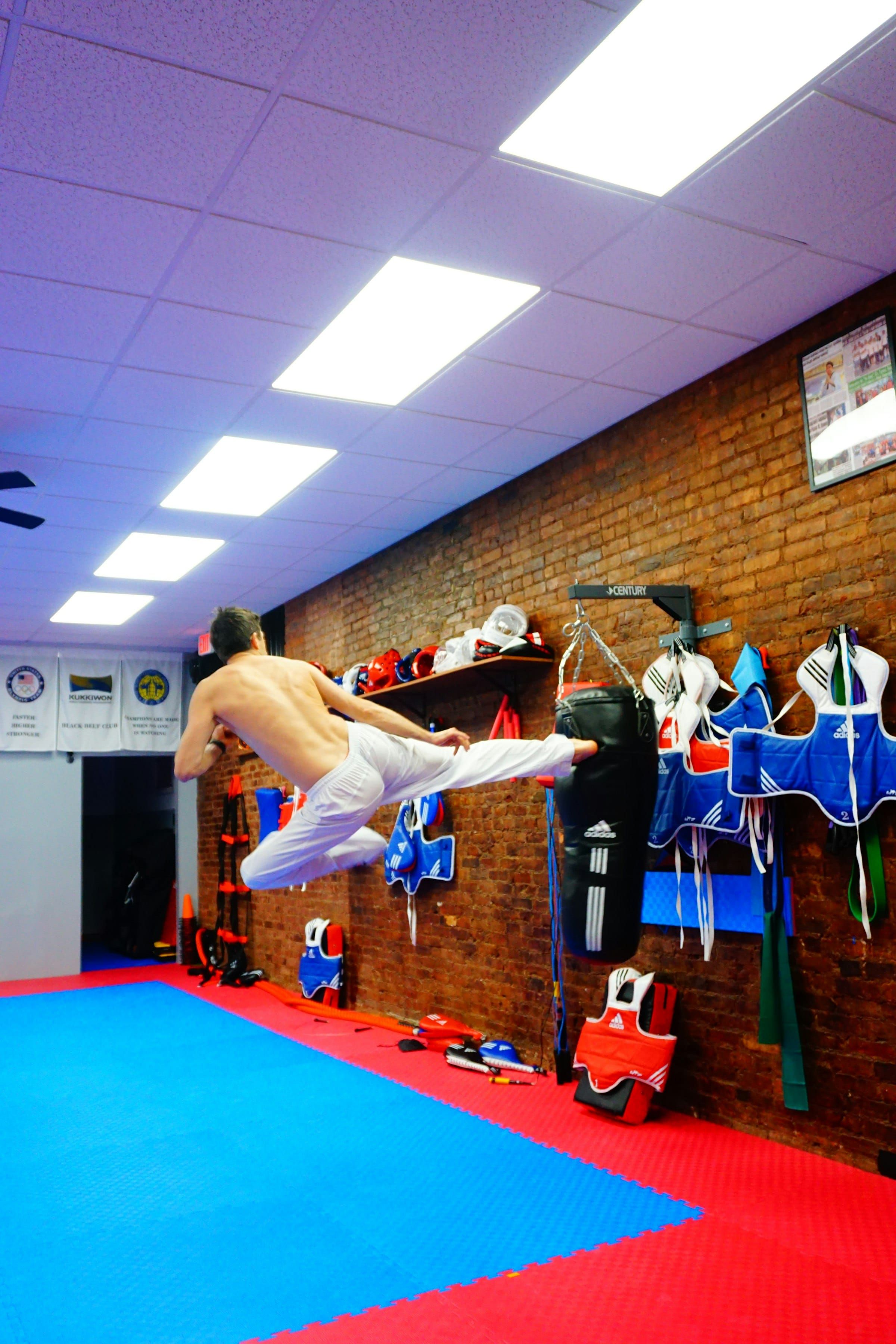 A shirtless man in white trousers throwing a high kick at a black heavy bag in a gym with a brick wall.