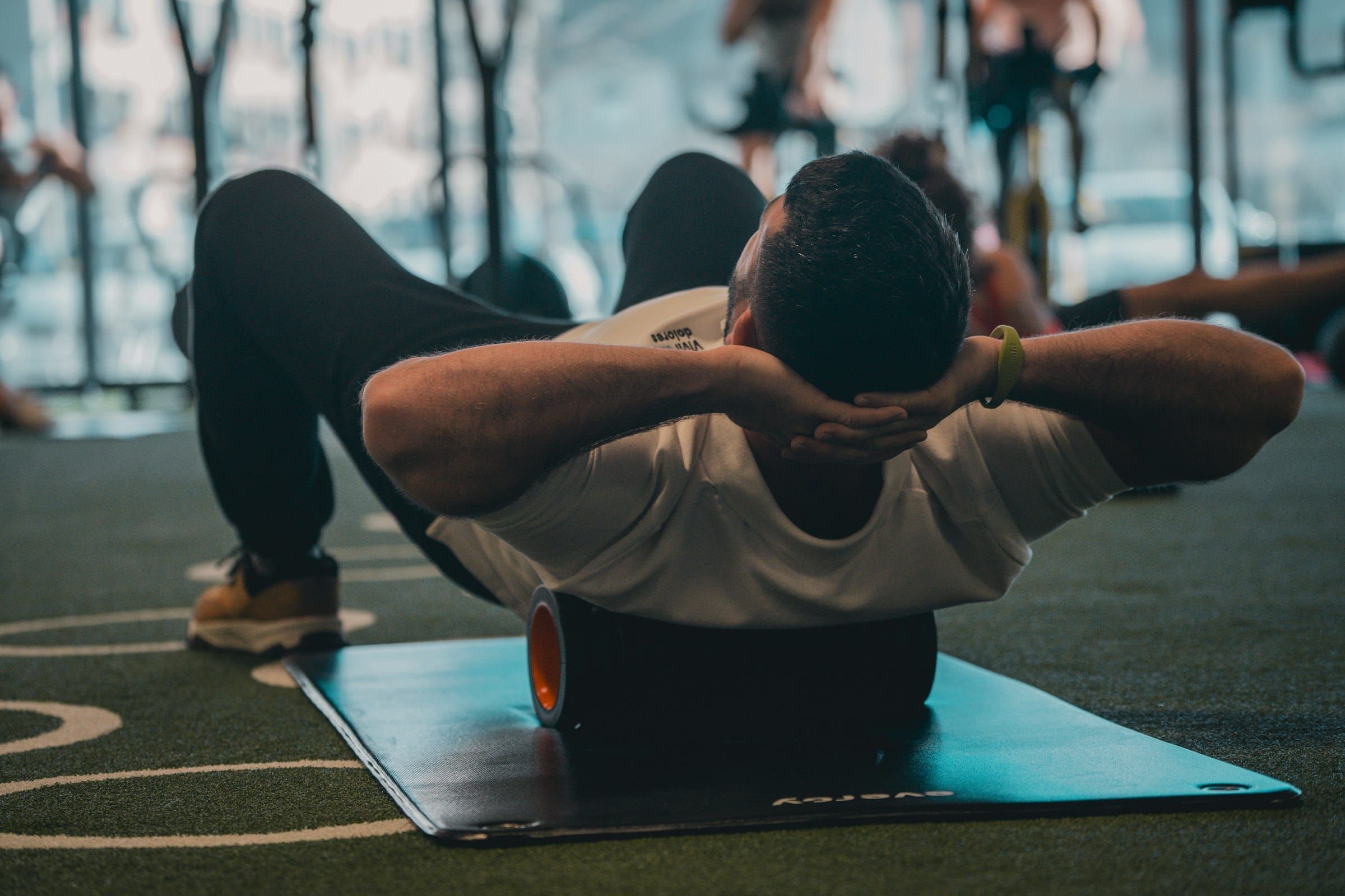 A man in a white T-shirt rolling his back over a foam roller on a mat in a gym.