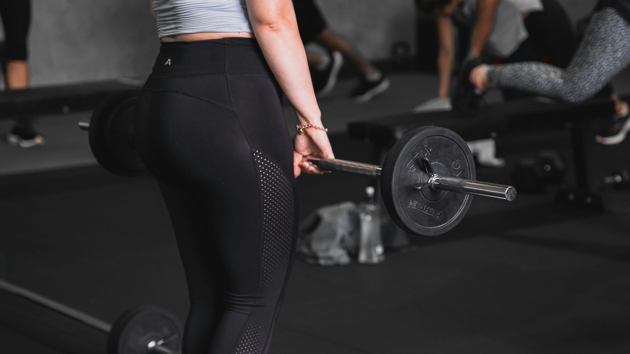 A woman in a grey crop top and black leggings lifting a loaded barbell in a dim gym, with other people training behind her.